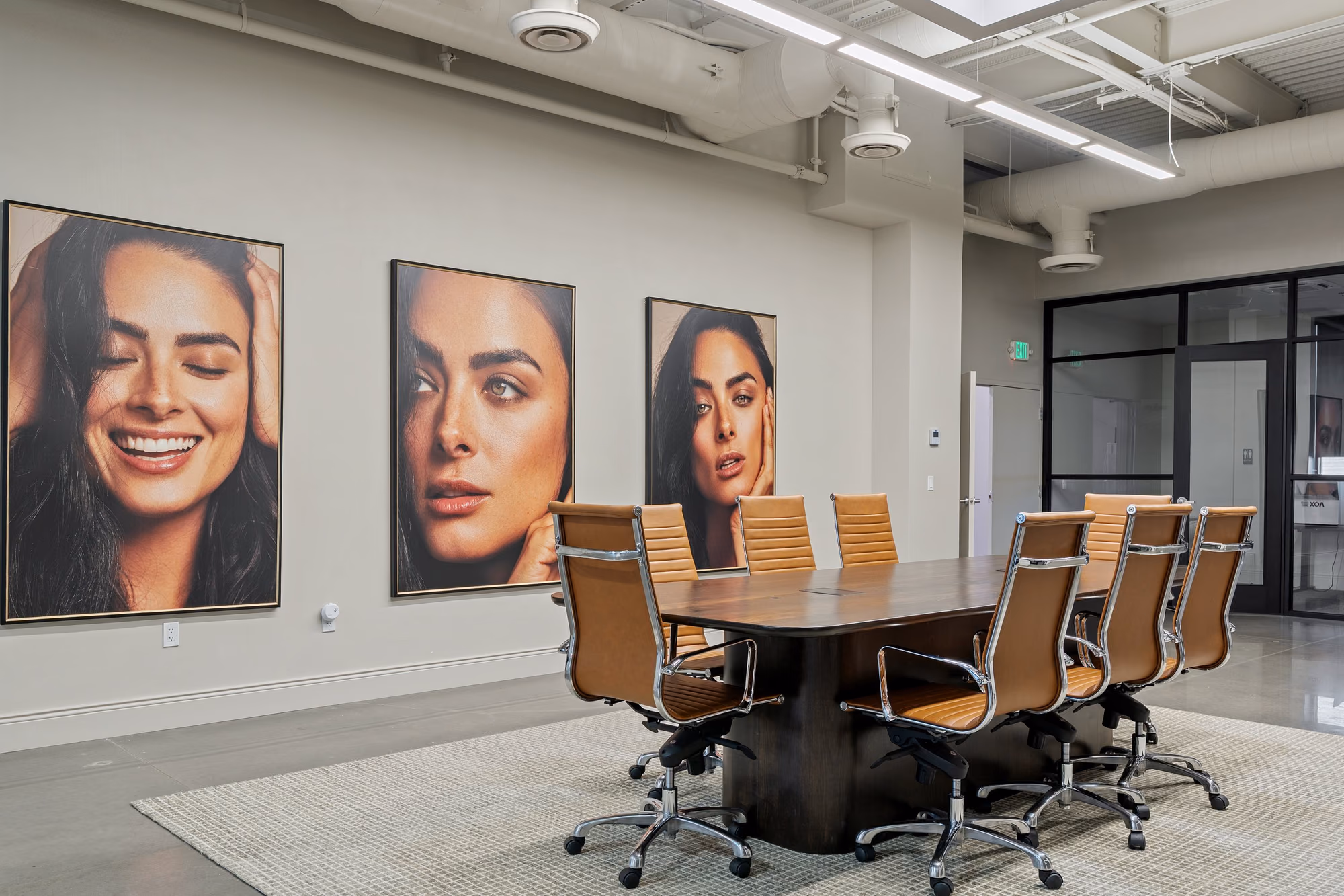Conference room with a dark wood oval table, tan leather office chairs, and three large framed portrait photographs on a grey wall, with exposed white ductwork overhead and a glazed corridor at right.