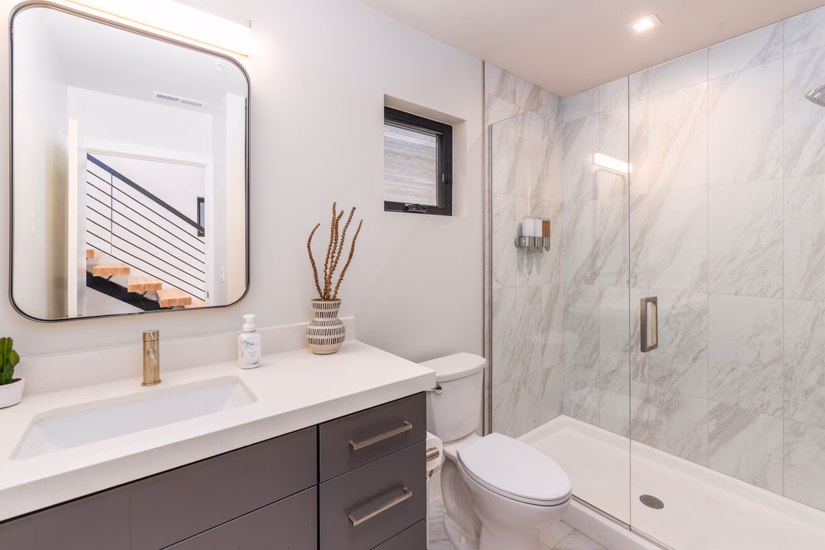 Bathroom with a floating dark vanity and white top, a rounded-corner mirror reflecting the stair, a marble-look tiled shower with a glass door, and a small window.