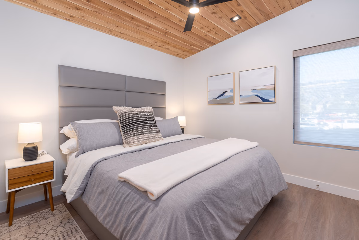 Bedroom with a cedar-plank ceiling, a grey paneled headboard, grey bedding, a wood nightstand with a lamp, two framed abstract prints, and a window.