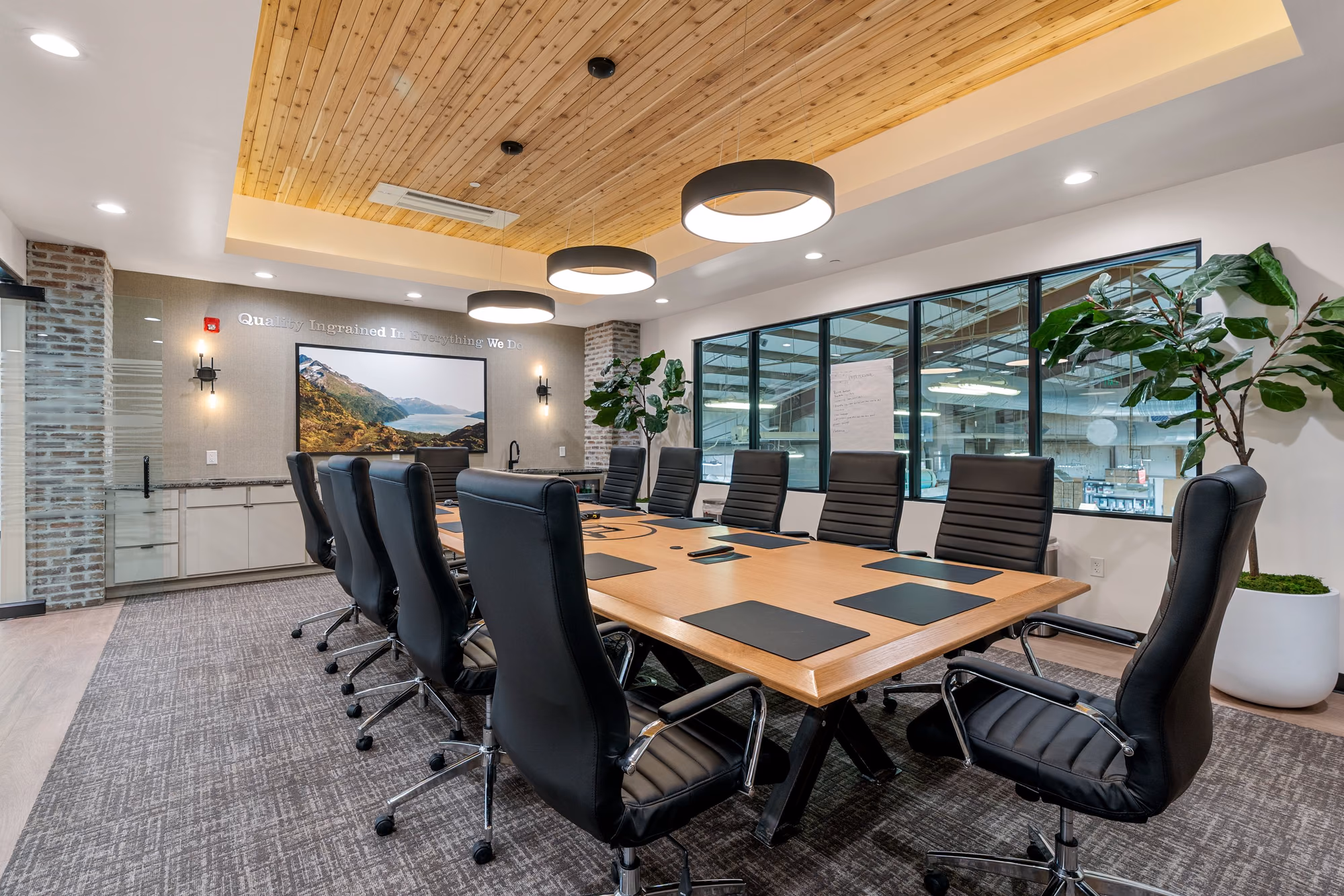 Boardroom with a recessed cedar-plank ceiling, round black pendants, an oak table, black leather chairs, a brick column, a landscape photograph, and windows onto the manufacturing floor.