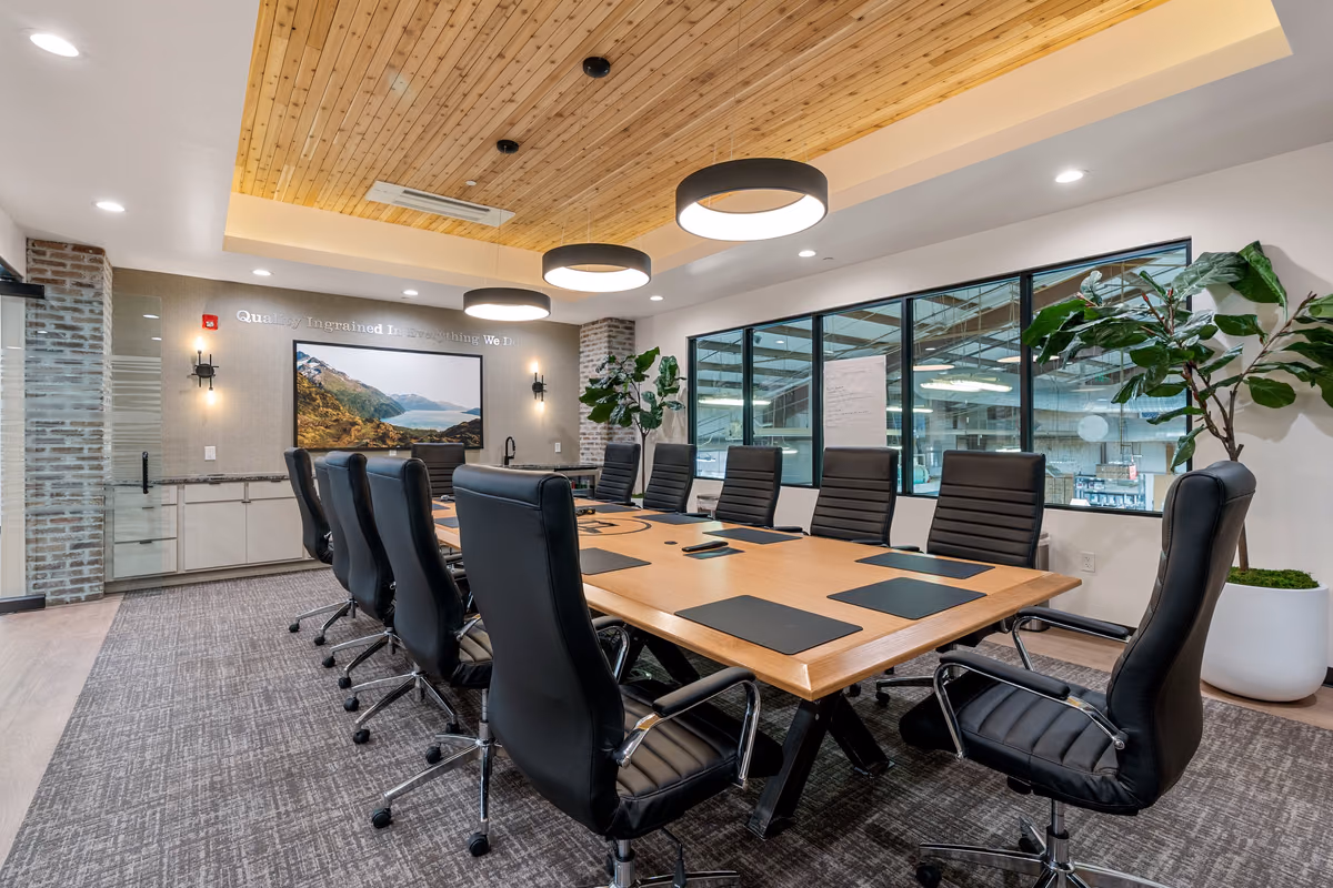 Boardroom with a recessed cedar-plank ceiling, round black pendants, an oak table, black leather chairs, a brick column, a landscape photograph, and windows onto the manufacturing floor.