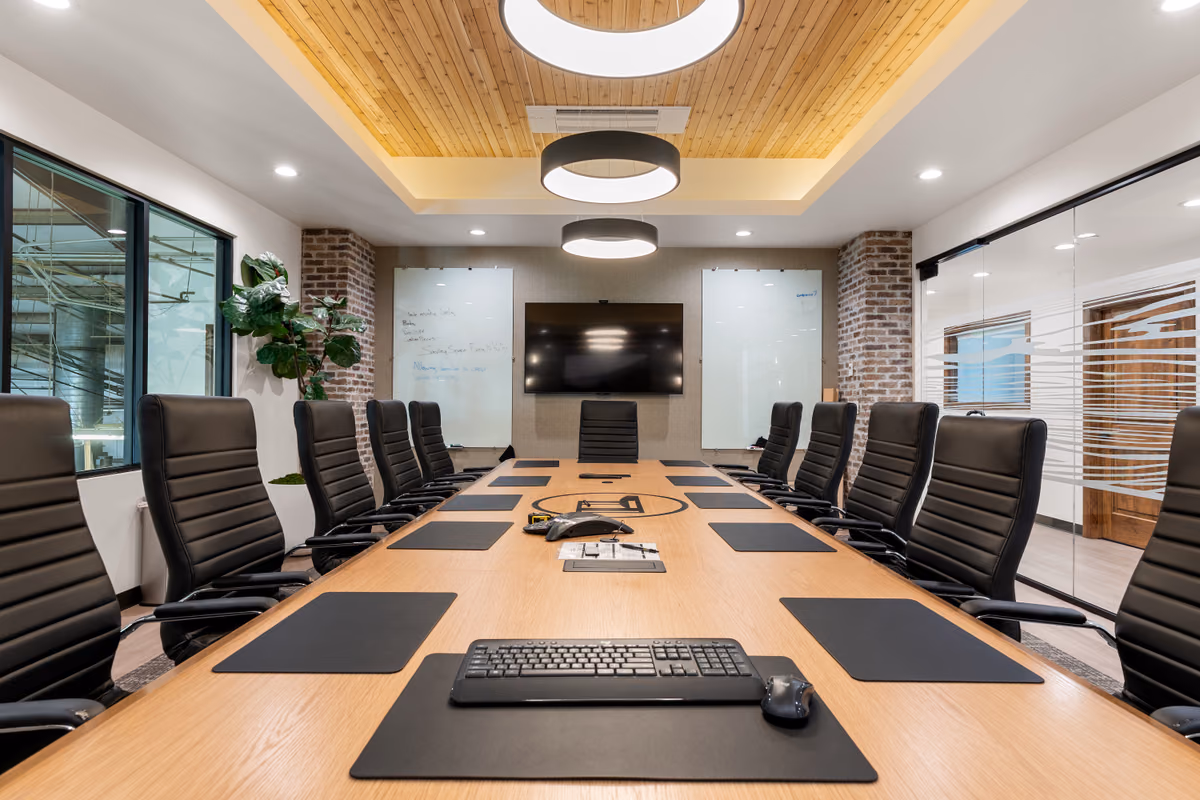 Boardroom seen down the length of an oak table with black leather chairs and desk pads, under a recessed cedar ceiling with round pendants, brick columns, and two glass whiteboards around a wall display.