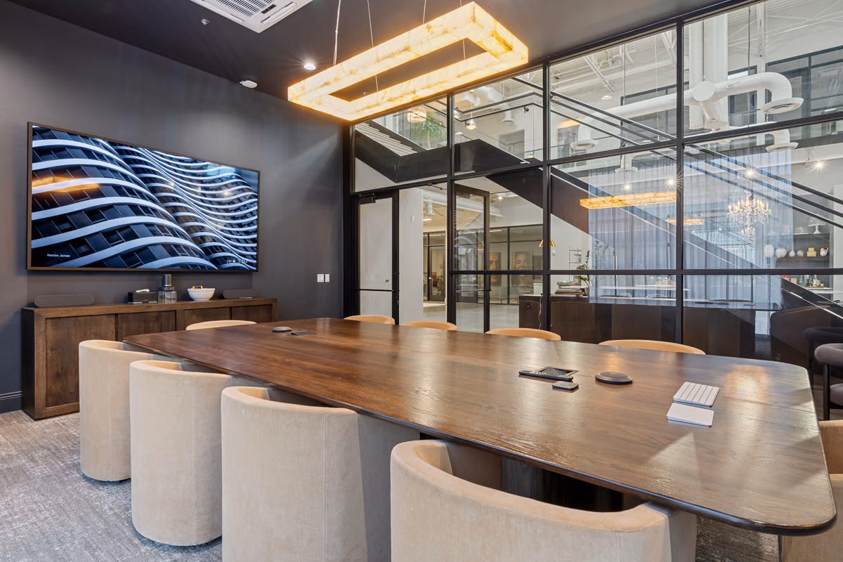Board room with a long dark wood table, cream upholstered chairs, a wall-mounted screen, a glowing rectangular pendant, and a glass wall with the stair and atrium visible beyond.