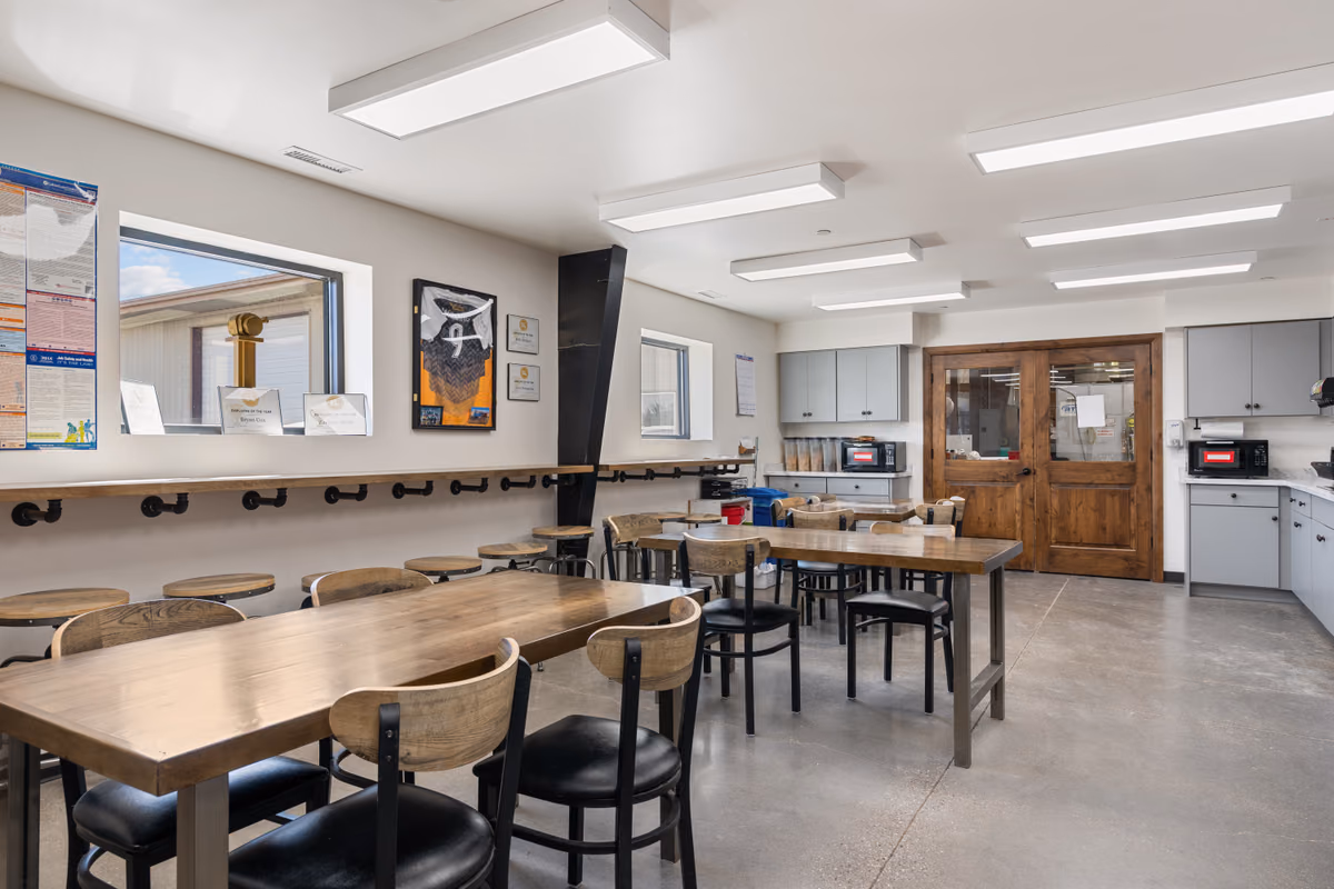 Employee break room with wood-topped tables, black chairs with wood backs, a pipe-bracketed counter with stools under a window, framed recognition plaques on the wall, and wooden double doors.