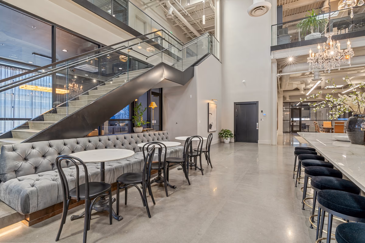 Double-height café with a grey tufted banquette, white marble bistro tables, black bentwood chairs, a glass-railed stair and mezzanine, a crystal chandelier, and a long counter with black stools.
