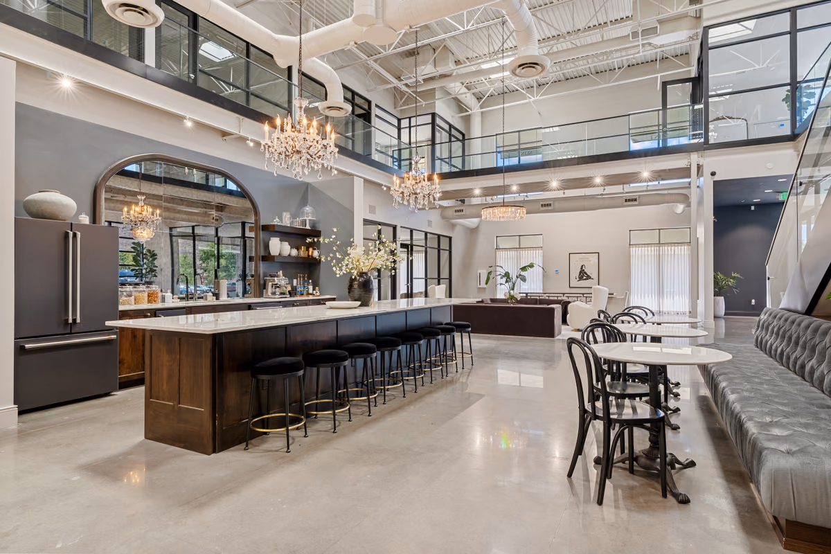 Wide view of the double-height café with a long dark wood island topped in white stone, black stools, two crystal chandeliers, an arched mirror behind a coffee counter, and the mezzanine above.