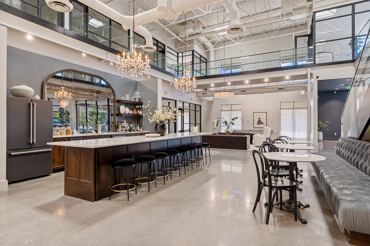 Wide view of the double-height café with a long dark wood island topped in white stone, black stools, two crystal chandeliers, an arched mirror behind a coffee counter, and the mezzanine above.