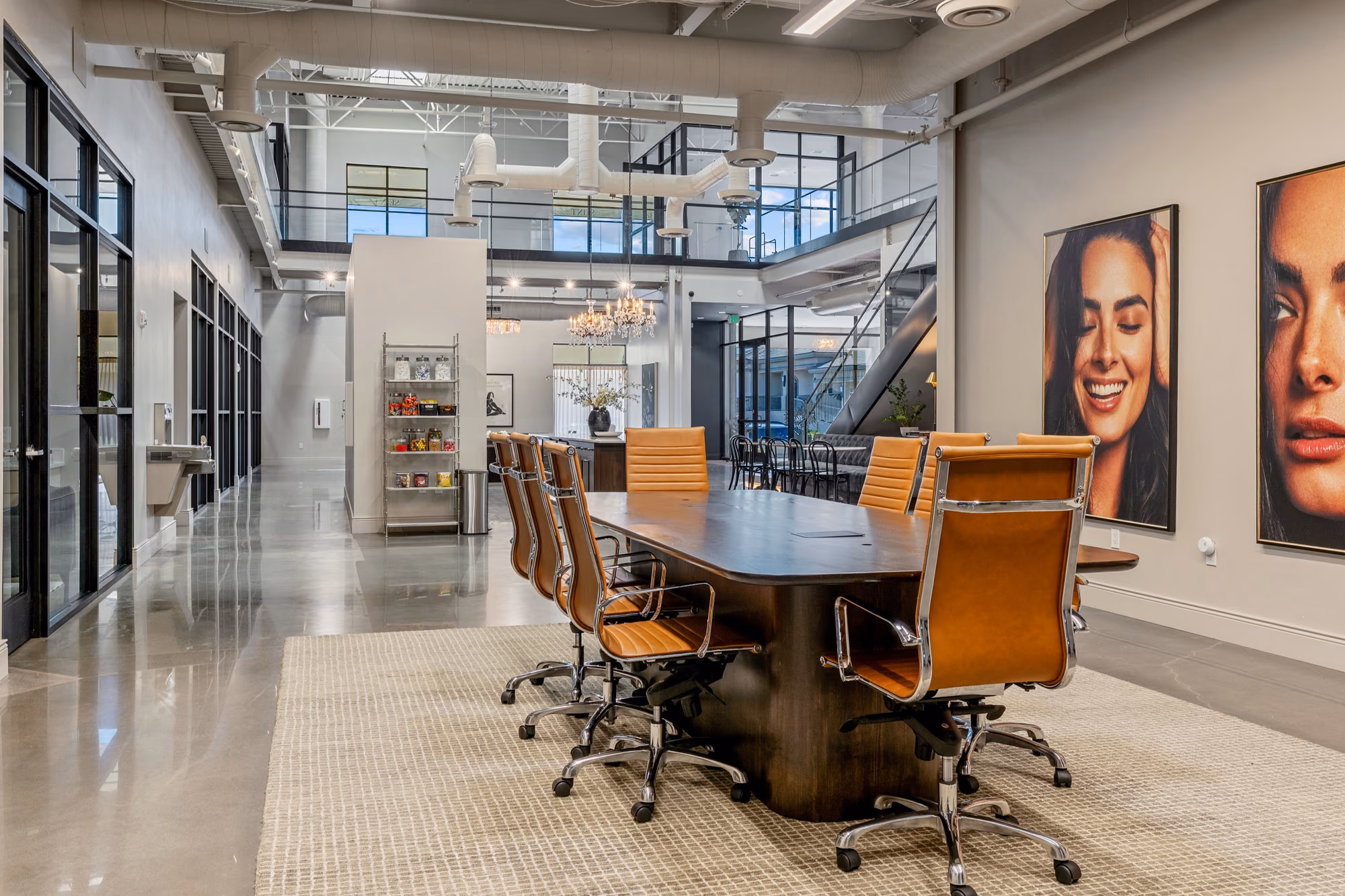 Open conference area with a dark wood table and tan leather chairs on a woven rug, large portrait photographs on the wall, a snack shelf, and a mezzanine with glass rails behind.