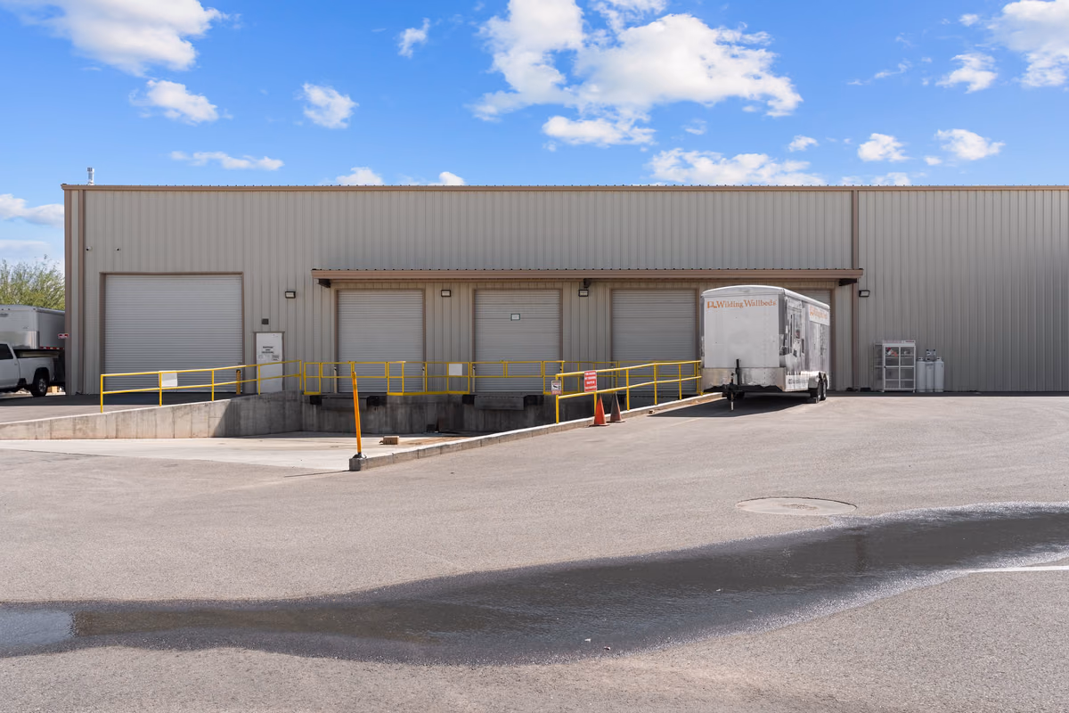 Rear of the manufacturing building with a row of roll-up loading dock doors, a yellow safety rail, a concrete ramp, and a Wilding Wallbeds trailer parked at one bay.