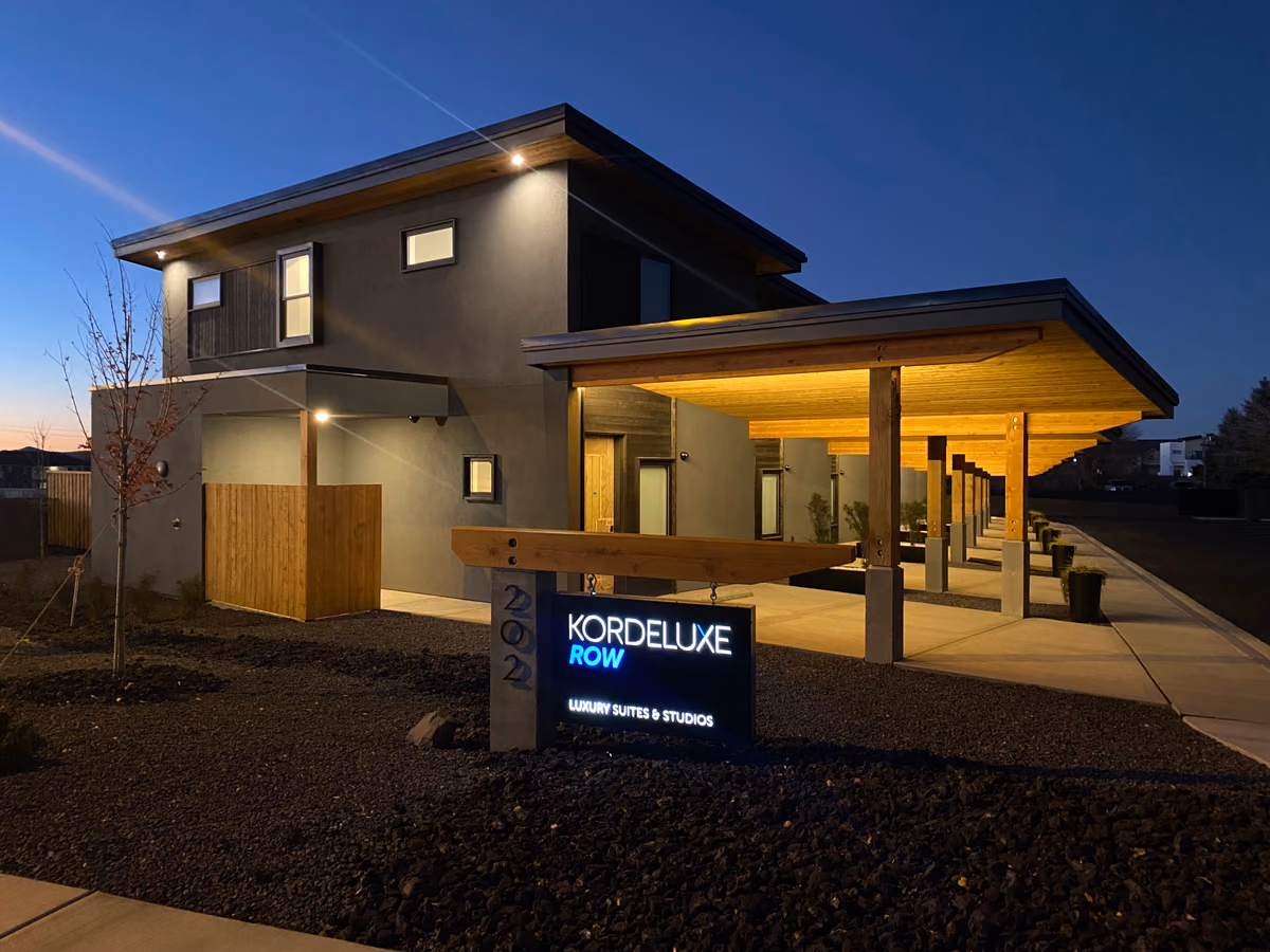 Two-story building at dusk with a shed roof, wood soffits, a long covered walkway on timber posts, and an illuminated monument sign reading Kordeluxe Row, Luxury Suites and Studios.