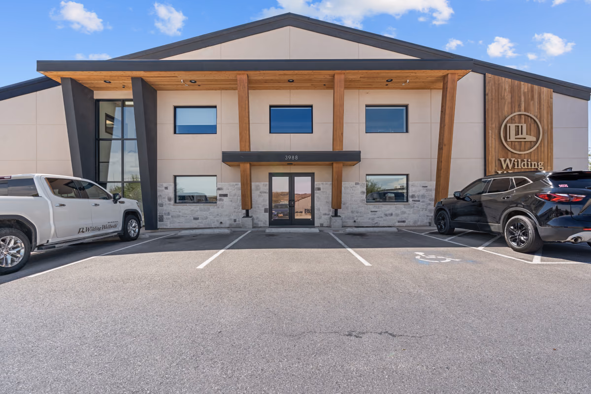 Front elevation of the building with cedar posts, stone base, and the sign wall, with a white pickup truck marked Wilding Wallbeds and a black SUV parked on either side.