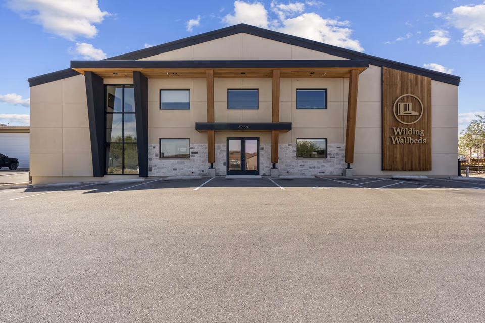 Symmetrical front elevation of a two-story building with a gabled roof, tapered black columns, cedar posts, a stone-veneer base, double glass entry doors, and a cedar sign wall reading Wilding Wallbeds, under a blue sky.