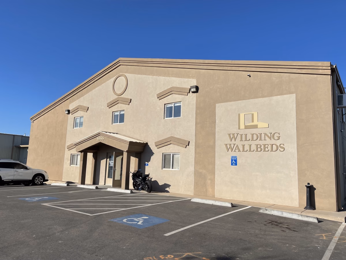 Three-quarter view of the building before renovation: tan stucco with chevron-shaped window awnings, a round window, a small gabled entry porch, and a Wilding Wallbeds sign panel.