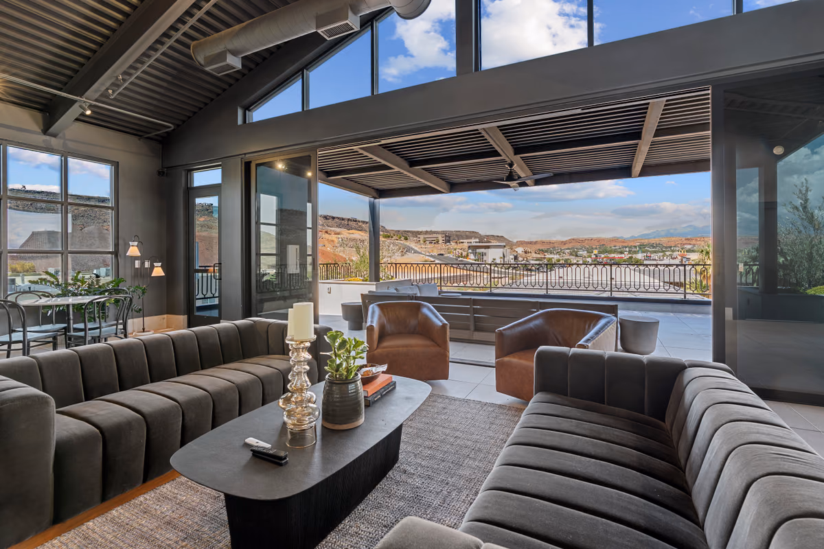 Rooftop lounge seating with grey channel-tufted sofas and leather chairs, looking through open glass sliding doors to a covered deck, an iron railing, and the city and red rock cliffs beyond.