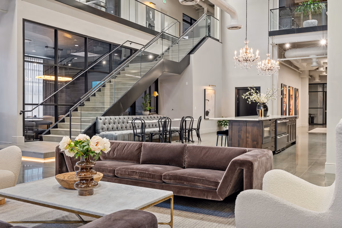 Close view of the lounge with a mauve velvet sofa, a marble-top coffee table with peonies, a glass and steel stair, a tufted banquette, chandeliers, and a dark wood kitchen island.