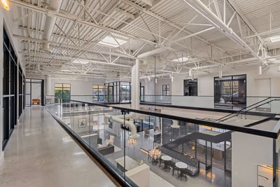 Mezzanine corridor with a polished concrete floor beside a glass guardrail, looking down on the café and banquette, under white steel trusses, skylights, and round ductwork.