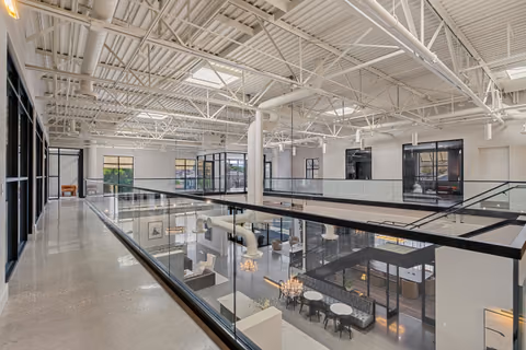 Mezzanine corridor with a polished concrete floor beside a glass guardrail, looking down on the café and banquette, under white steel trusses, skylights, and round ductwork.