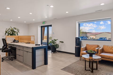 Front office with a light stone-topped desk with a navy frame, a telephone, a monstera plant, tan leather armchairs, a live-edge coffee table, and a window looking out to a neighboring building and hills.