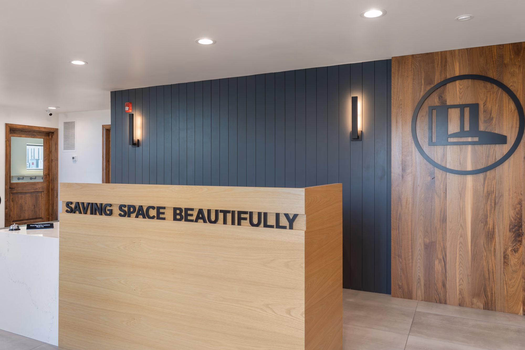 Reception with a light oak front desk carrying black lettering that reads Saving Space Beautifully, a dark slat wall with two wall lights, a walnut panel with the company logo, and a stone counter at left.