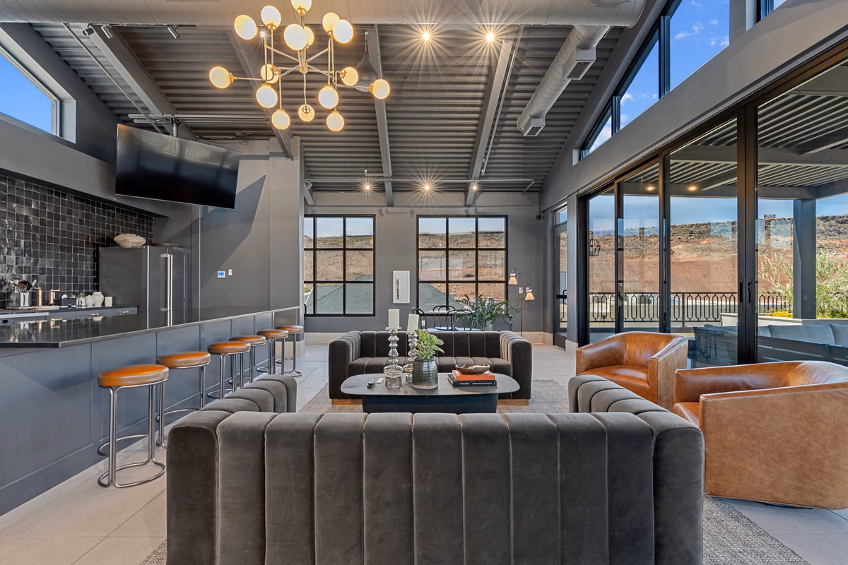 Rooftop lounge with a counter of tan leather bar stools at left, a black-tiled wall, globe chandeliers under a corrugated ceiling, grey velvet sofas, and sliding glass doors to the deck.