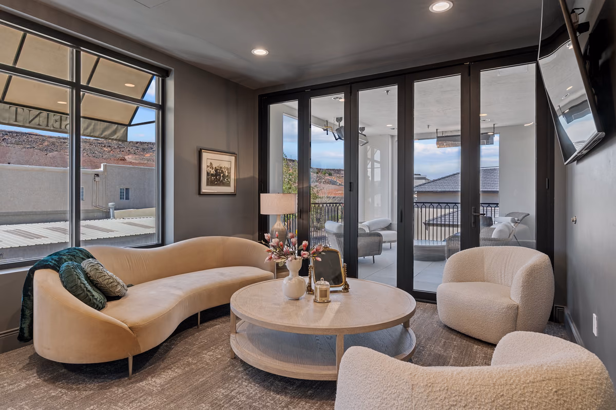 Sitting room with a curved cream sofa, a round pale wood coffee table, white boucle chairs, a wall-mounted television, and folding glass doors opening to a covered balcony.