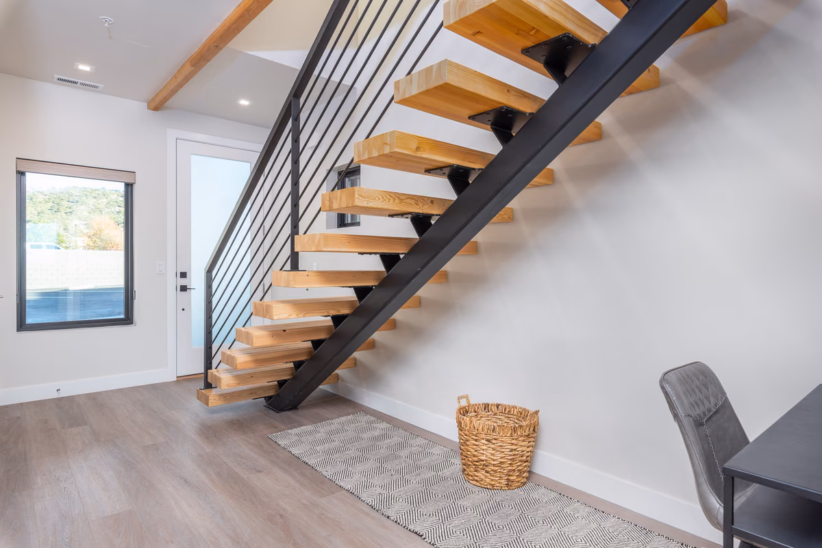Timber-tread stair with a black steel stringer and rod railing, a glazed front door, a window, a basket on a patterned runner, and a desk and chair at right.