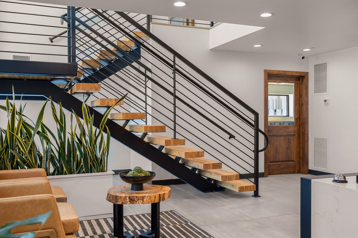 Lobby stair with solid timber treads on a black steel stringer and horizontal rod railing, beside a planter of snake plants, a live-edge side table, and tan leather chairs.
